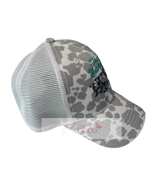 side view. “Women’s gray and white cow‑print baseball hat featuring green ‘Thank a Farmer’ embroidery above a detailed black stitched cow graphic on the front.