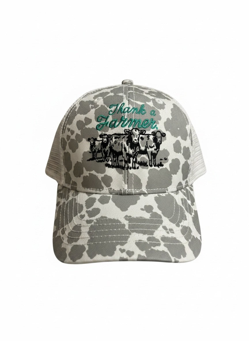 Gray and white cow‑print baseball hat with green embroidered ‘Thank a Farmer’ lettering and detailed black stitched cows on the front panel.