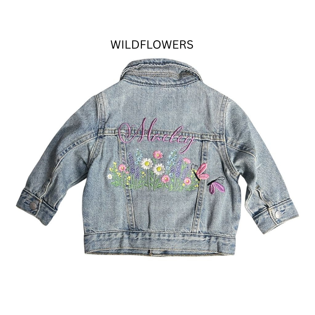 Personalized light blue denim jacket with pink embroidered name ‘Moxley’ above detailed floral embroidery, green grass, and pink and purple butterflies on the back panel.